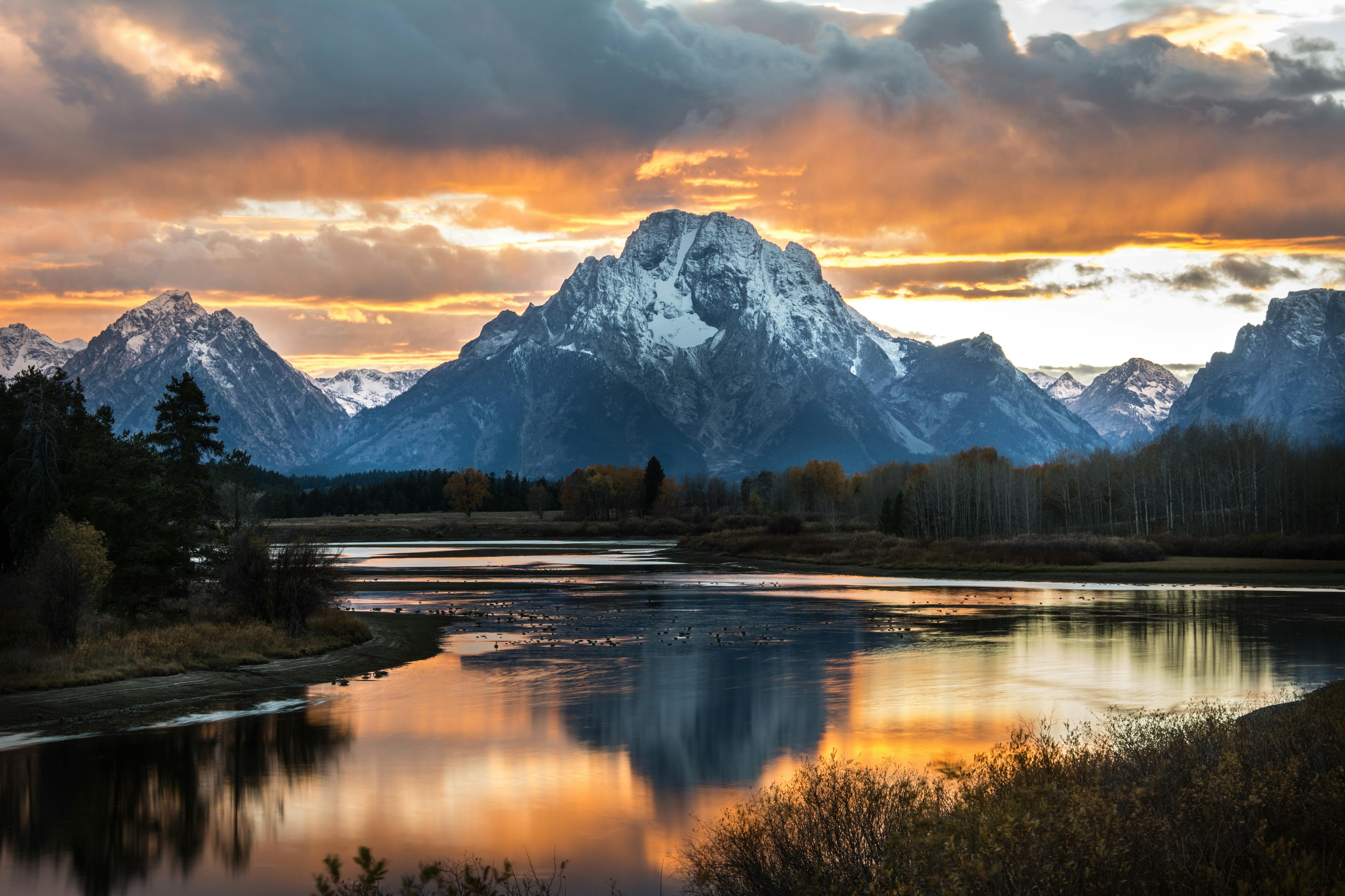The Tetons rise in the background of the image with the sun setting behind them. The sunset is reflected on a wide river in the foreground.