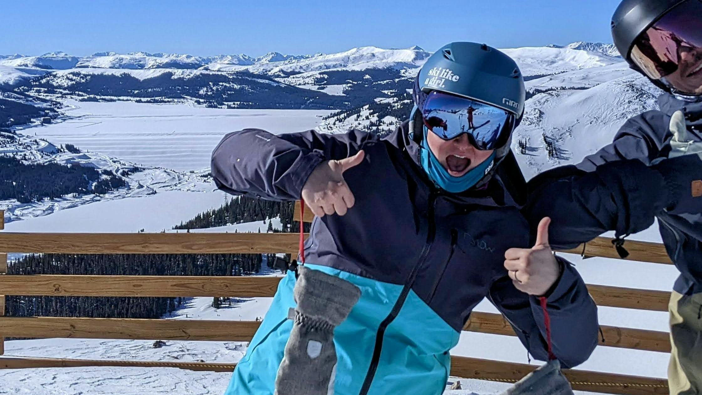 Two skiers on a ski run giving a thumbs up to the camera. There are snowy mountains in the background. 