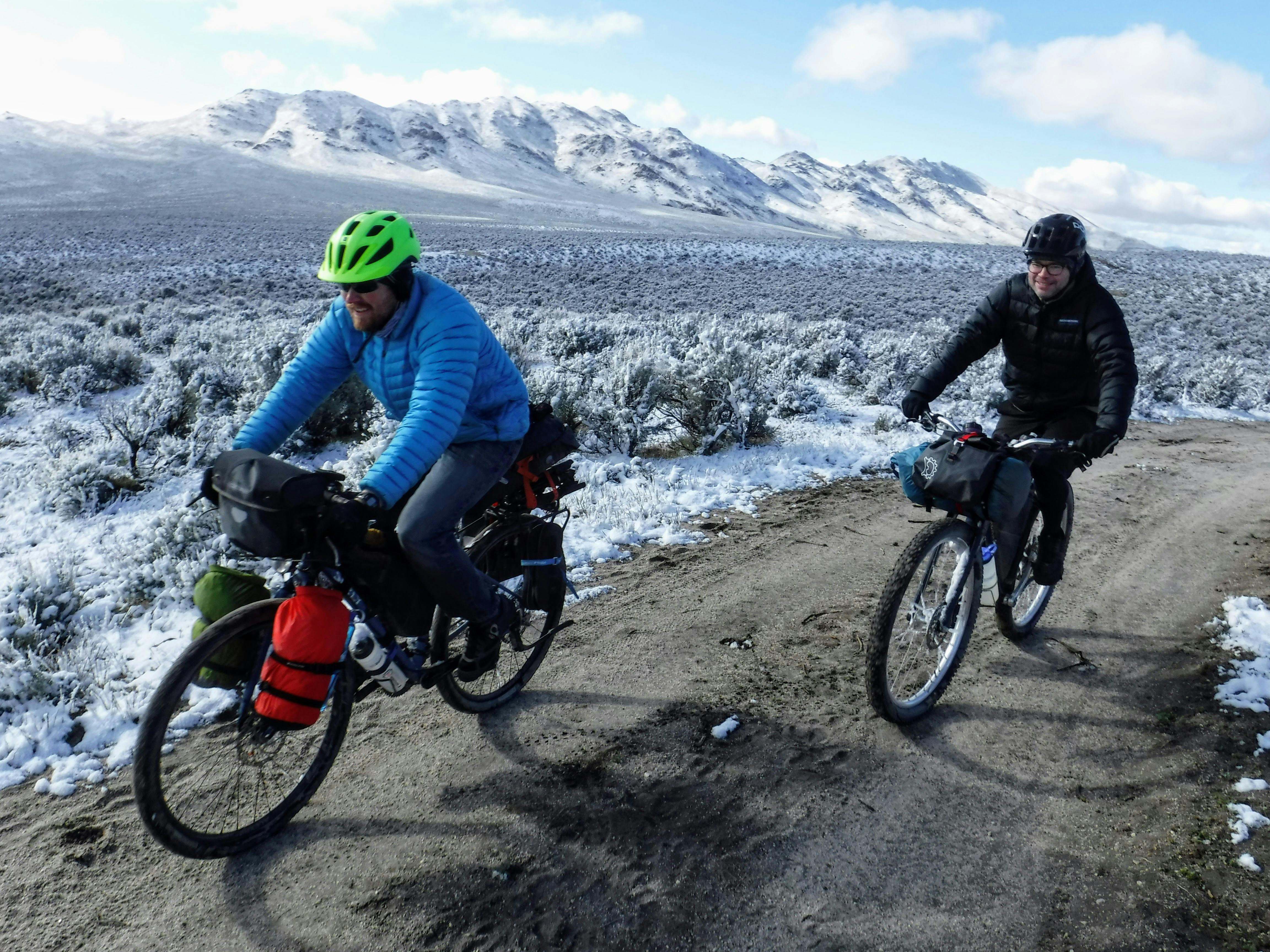 Two bikepackers wear their puffy jackers as they ride along a trail. There is snow on the ground on the sides of the trail.