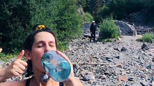 A woman drinks water out of a blue plastic bottle while giving the camera a thumbs up.