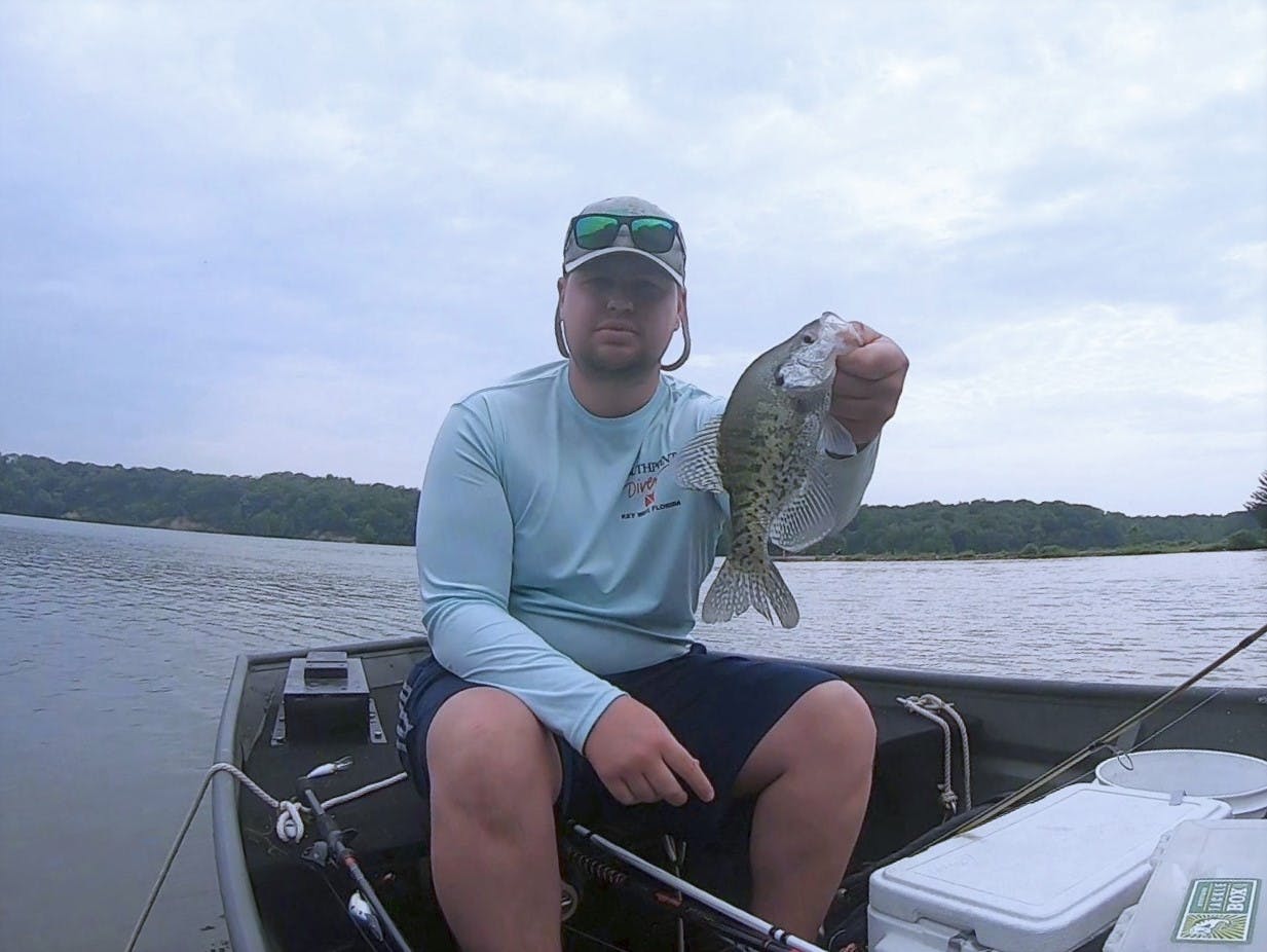 A man sits on a boat and holds out a freshly-caught crappie to the camera.