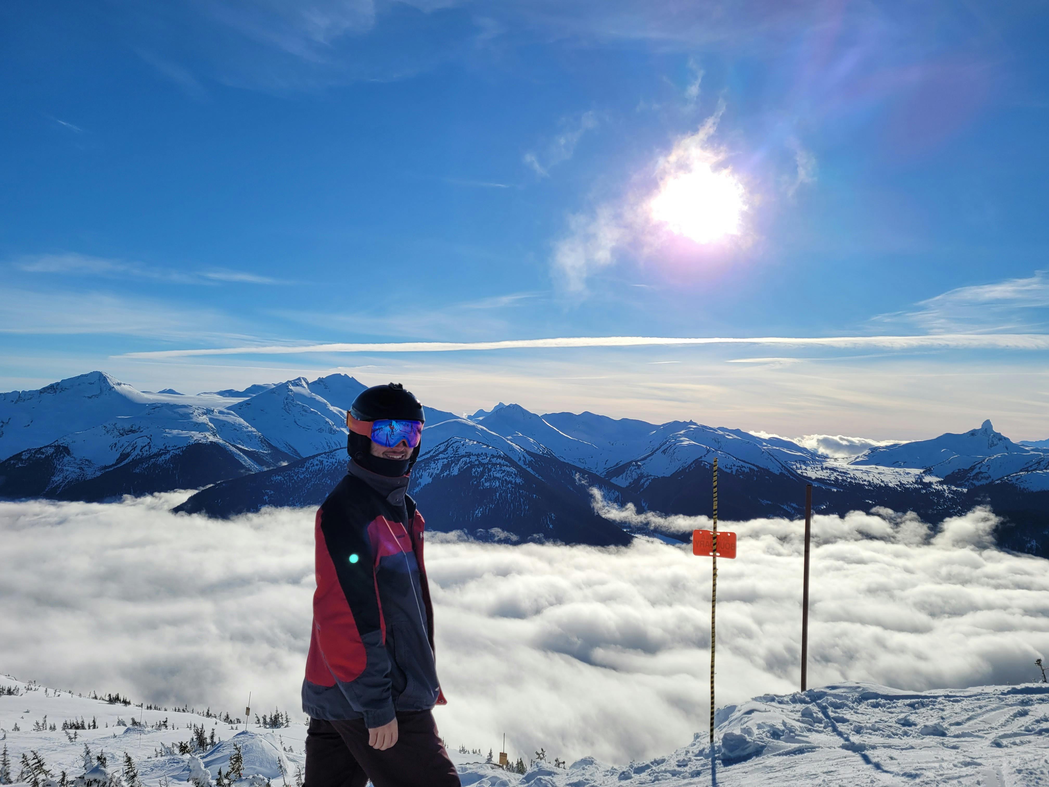 A skier at the top of a snowy peak with clouds and snowy mountains in the background. 