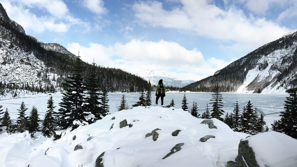 A person stands on a snowy hill looking out at a lake in winter