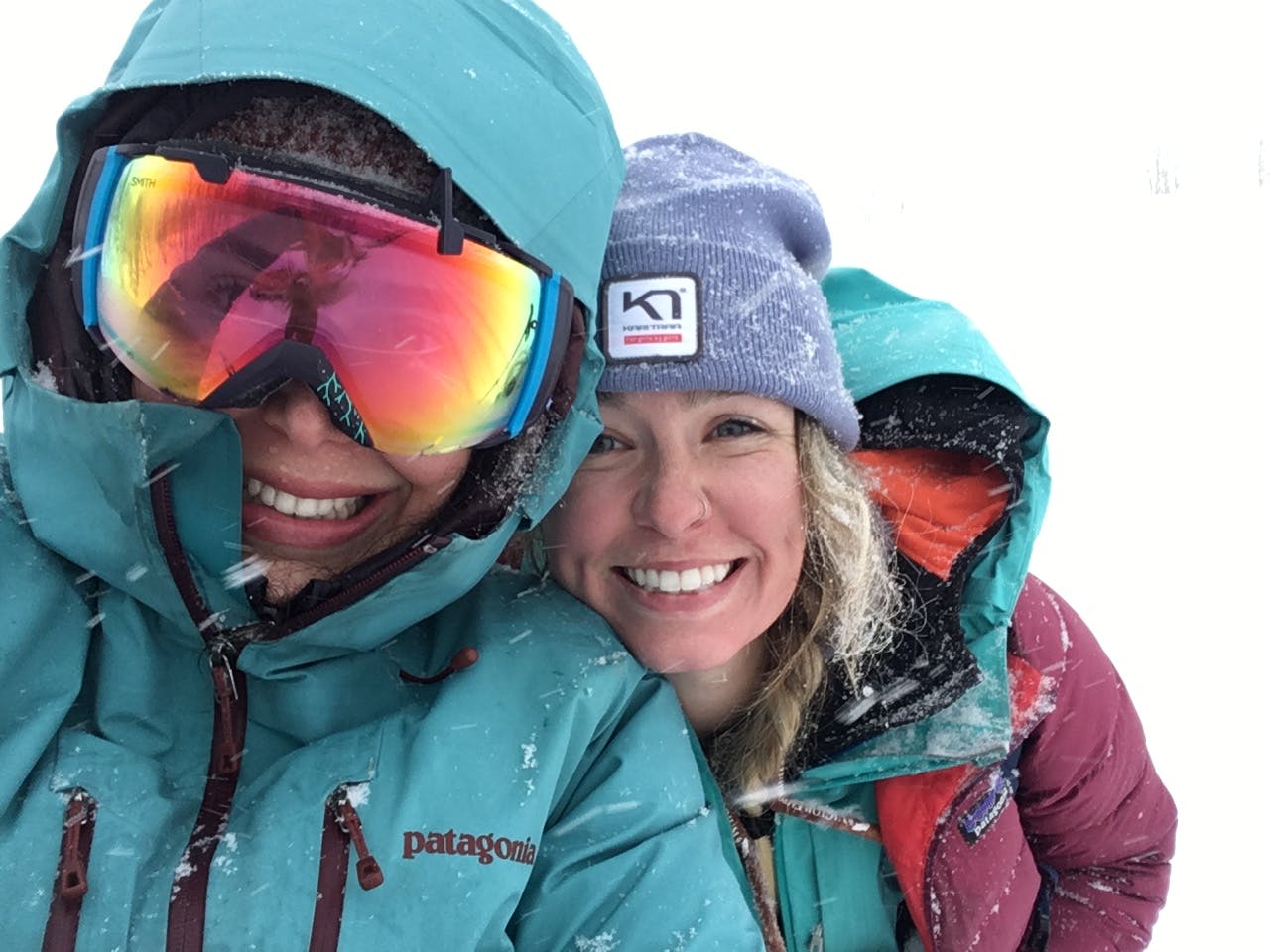 Two people smiling in the snow. One is wearing goggles and the other is wearing a Patagonia Hi Loft Jacket. 