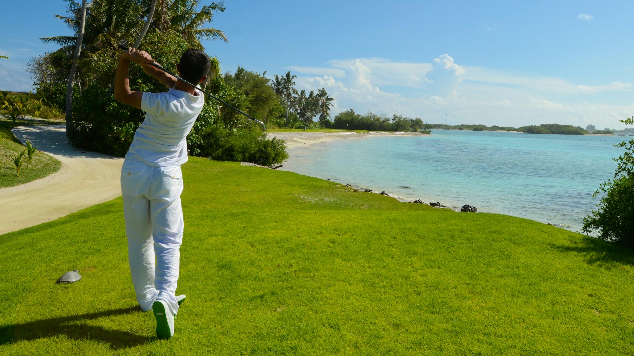A man swings a golf club on a course next to tropical waters
