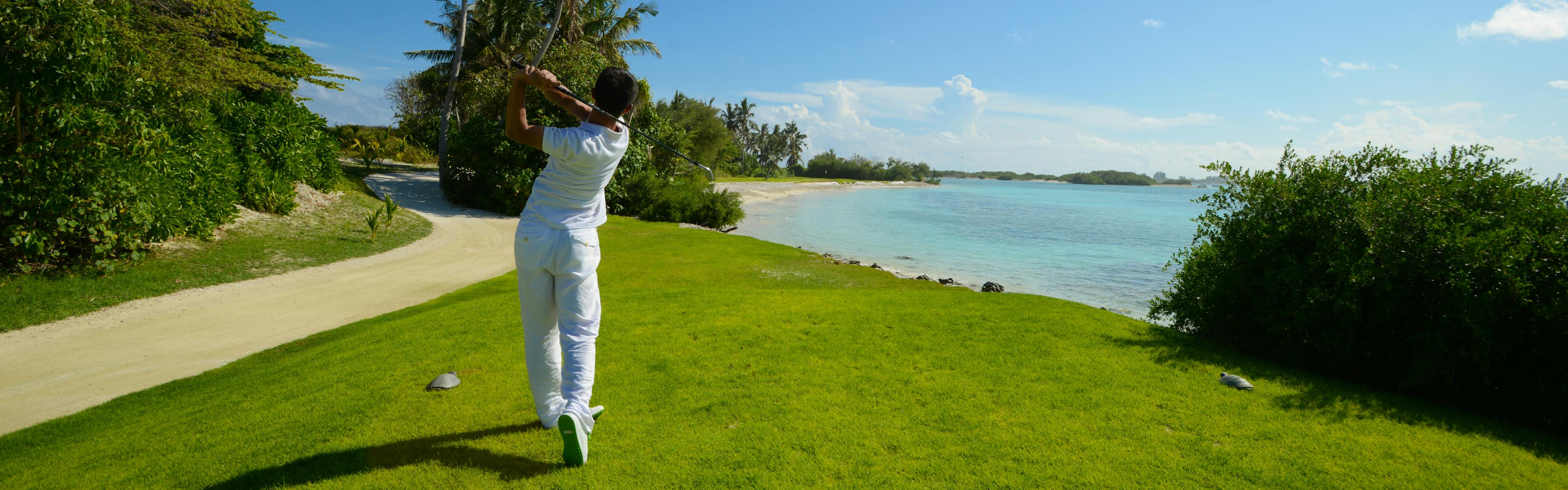 A man swings a golf club on a course next to tropical waters