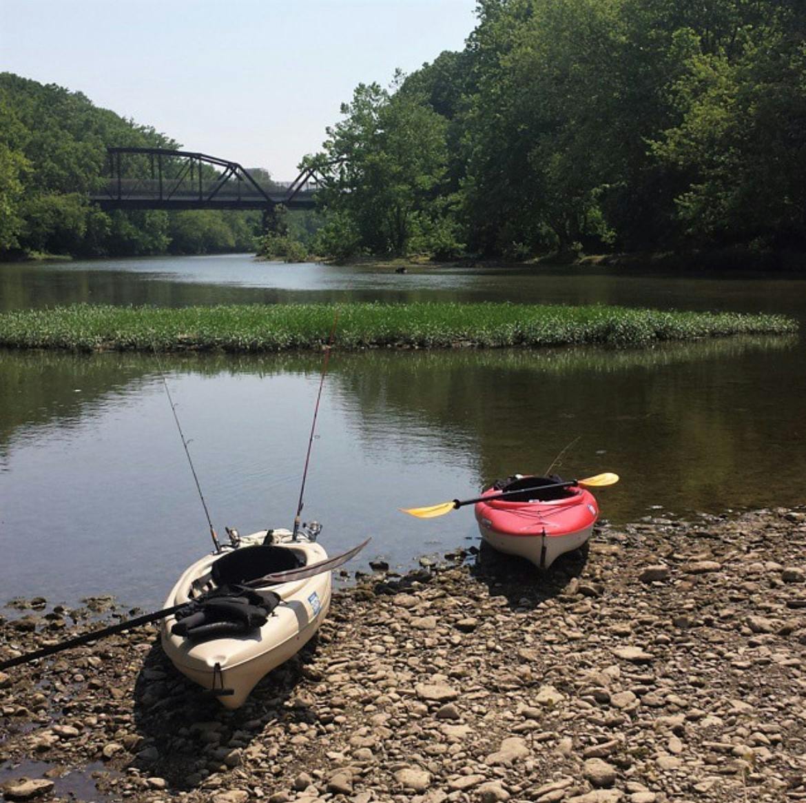 Two kayaks sit on the bank of a river. One is red and one is tan.