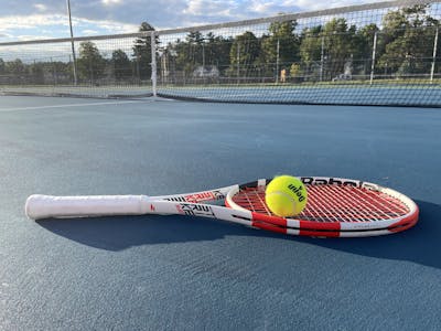The Babolat Pure Strike 18x20 Racquet lying on the tennis court with a tennis ball.