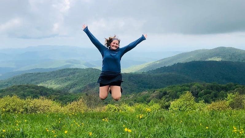 The author jumps in the air with her arms outstretched and her knees tucked under her. The grass is lush and green and dotted with yellow wildflowers. There are rolling hills in the background. 