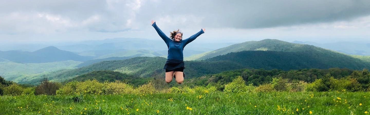 The author jumps in the air with her arms outstretched and her knees tucked under her. The grass is lush and green and dotted with yellow wildflowers. There are rolling hills in the background. 