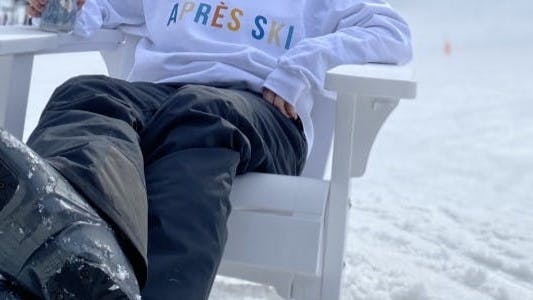 A woman in ski boots and ski pants sits in a chair. There is snow behind her.
