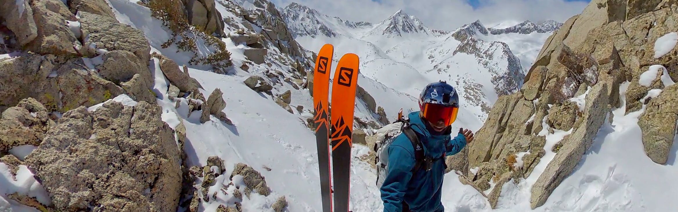 Josh takes a selfie on the mountain while wearing a google and a helmet. His Salomon skis are propped up next to him.