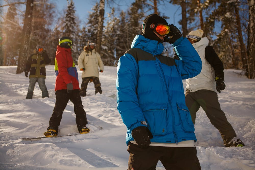 Five snowboarders stand on a hill. The one in front is wearing a blue jacket and adjusting his goggles. 