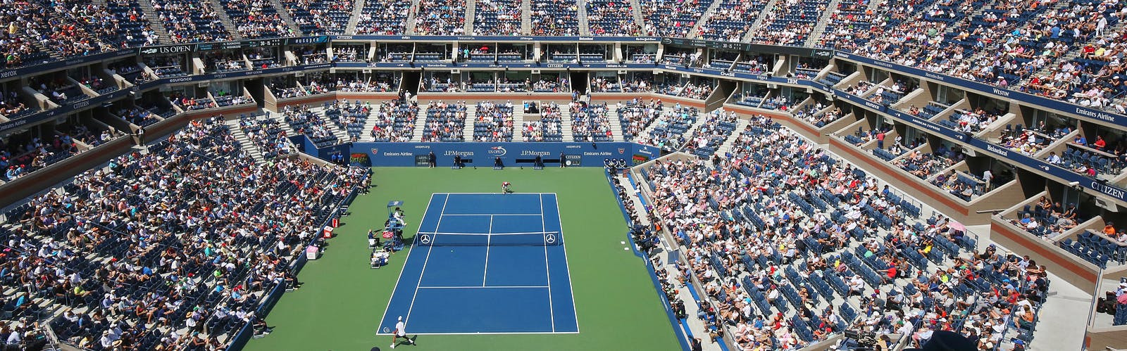 Arthur Ashe Stadium during the day, filled with fans watching a men's singles match.