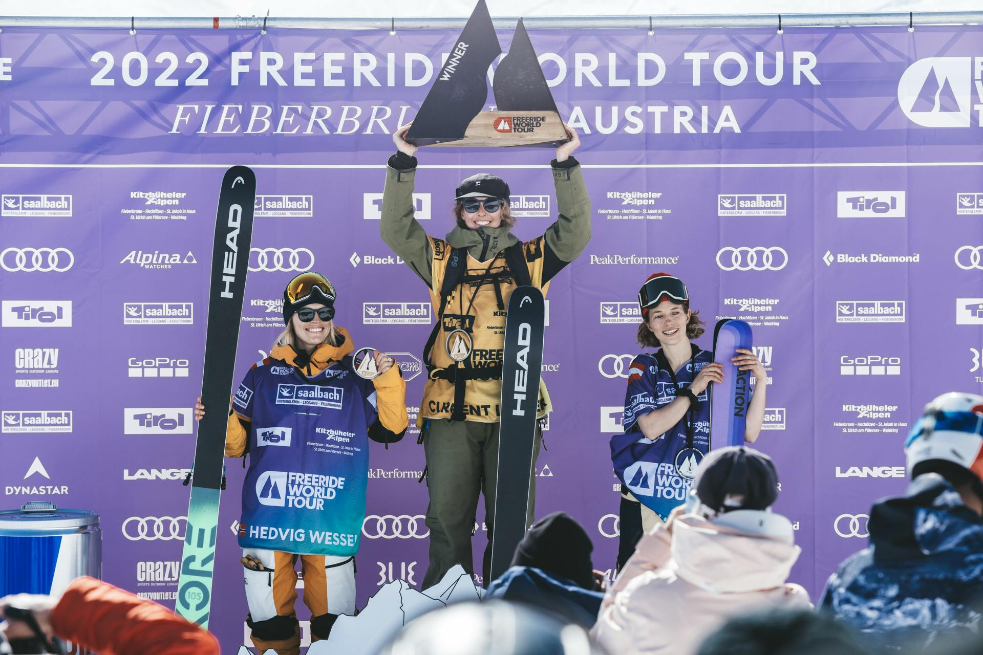 The top three female skiers of Stop Four of the FWT stand on the podium in front of a purple backdrop.