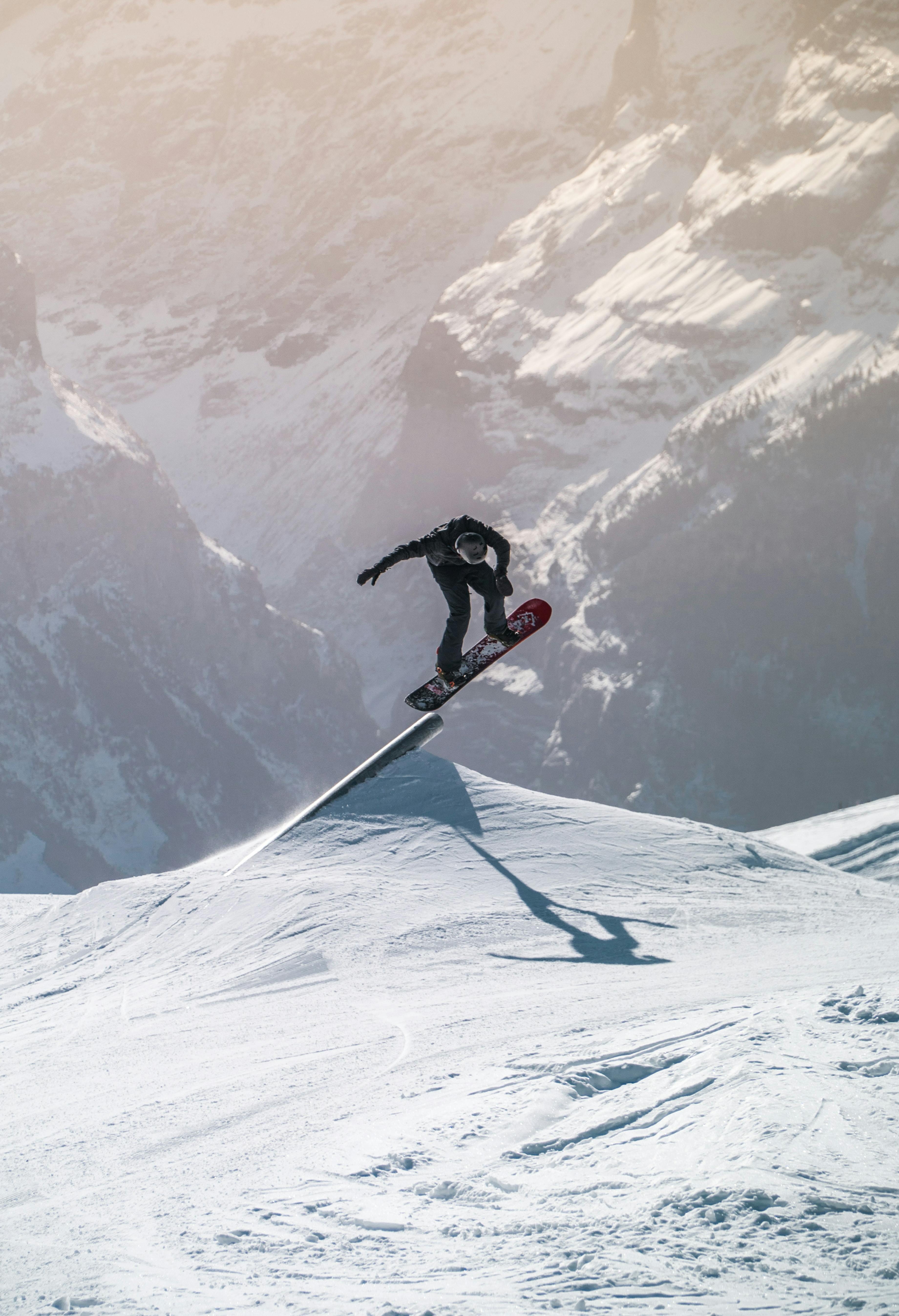 Snowboarder wearing all black slides off of a terrain park feature that is part rail, part jump on his snowboard. Dramatic mountains are in the background.