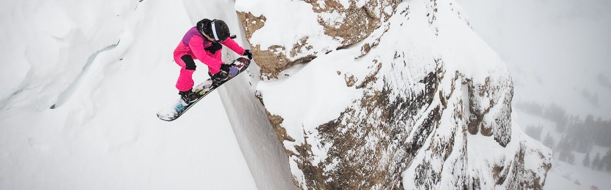 A snowboarder in pink sits at the lip of the coulier. 