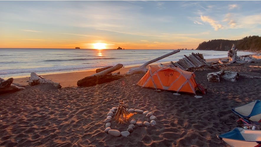 A tent and a campfire set out on a beach with the sun setting into the Pacific.