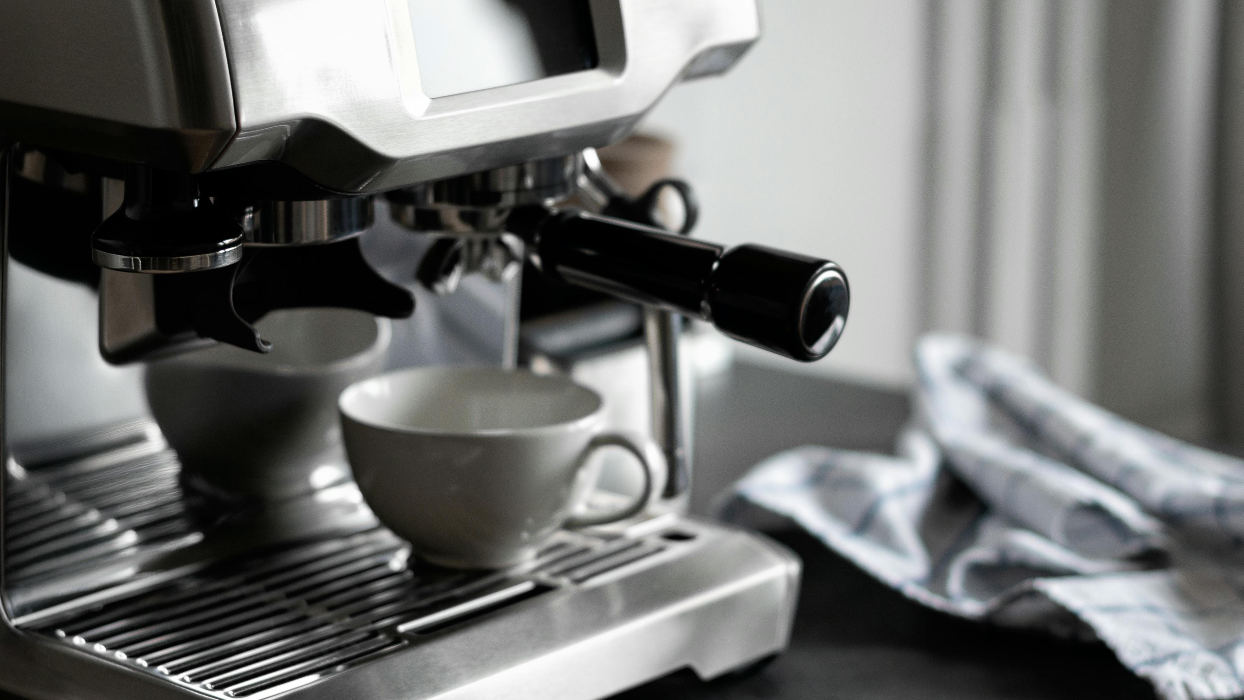 A white espresso cup sits on a stainless steel espresso machine, waiting for the shot. 