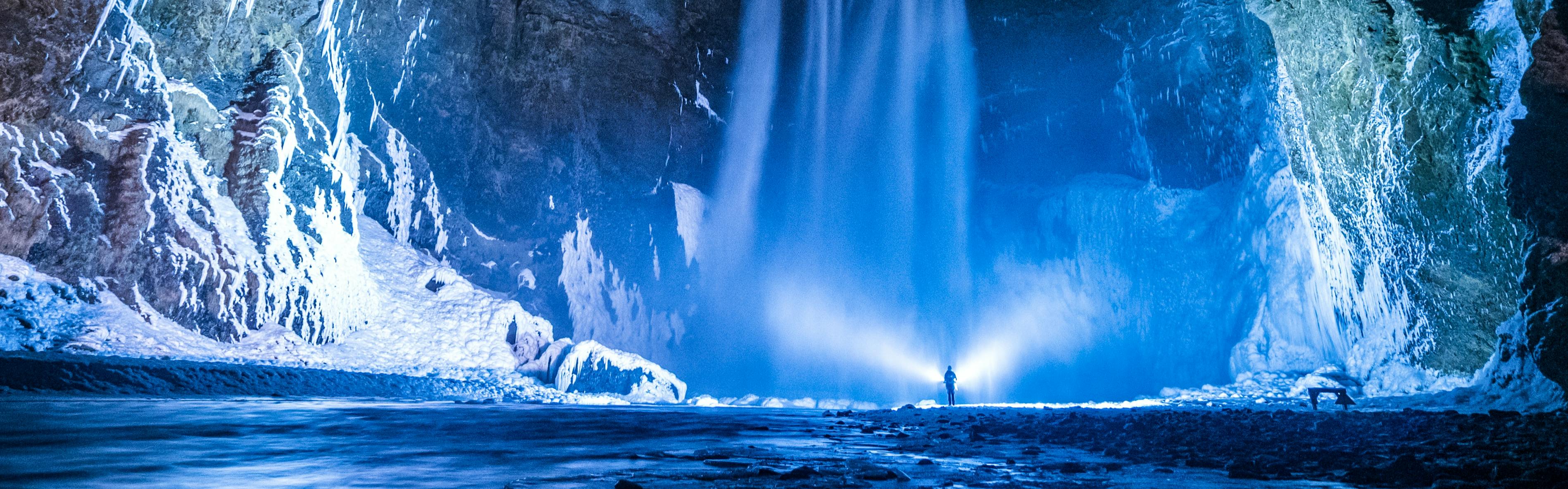 A dramatically lit waterfall at night, a person standing in front of the falls in the distance