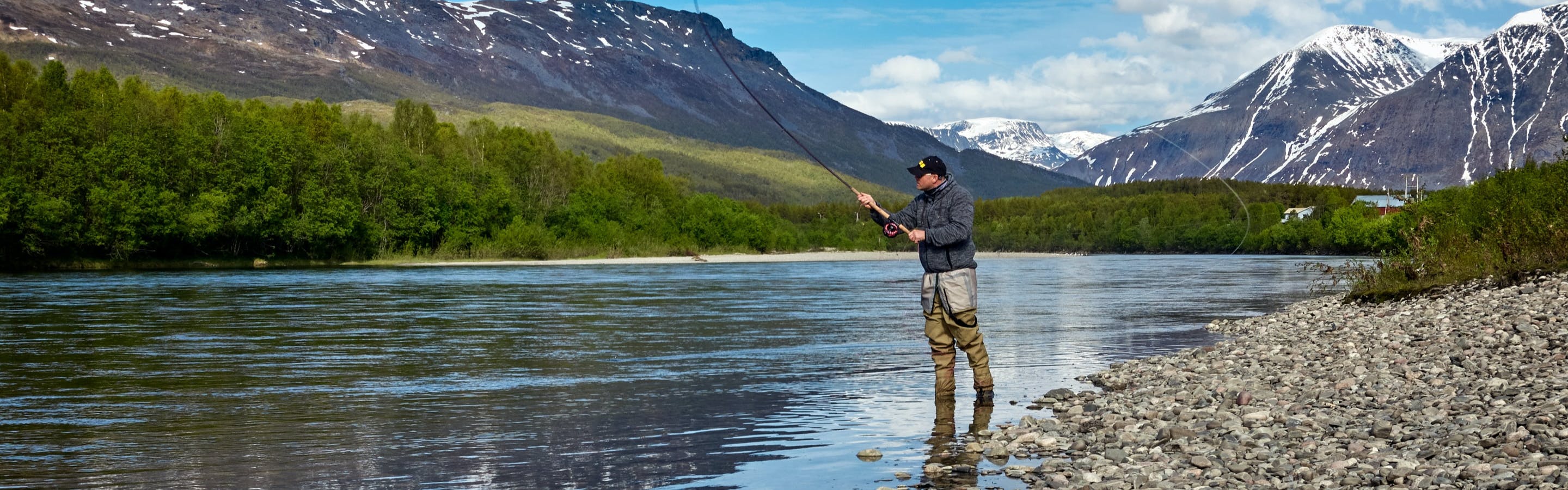 A fly fisherman standing in the river near the bank casting out a fly with snow-topped mountains in the distance