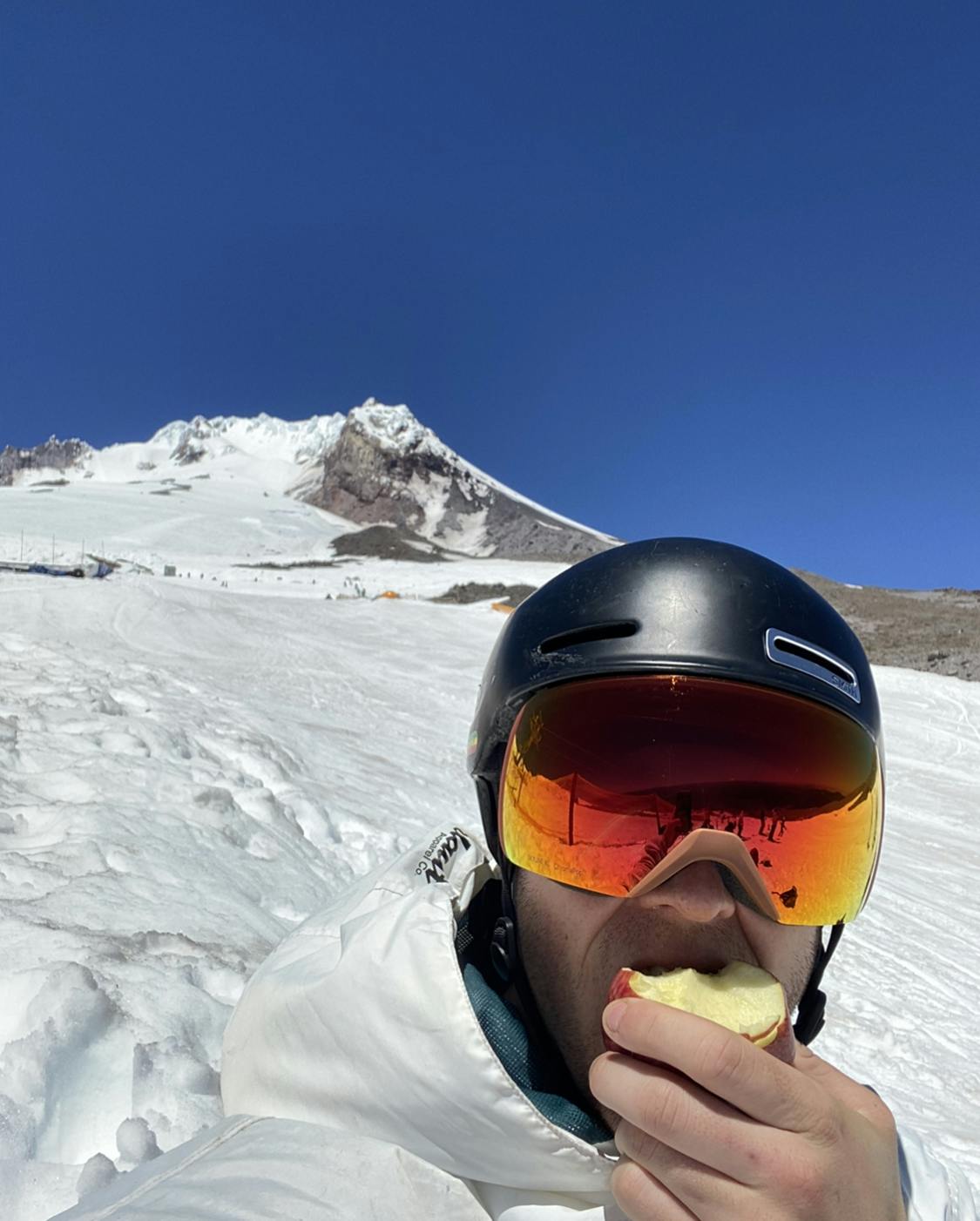 A skier wearing the Smith Skyline XL goggles and eating an apple on a snowy mountain. 