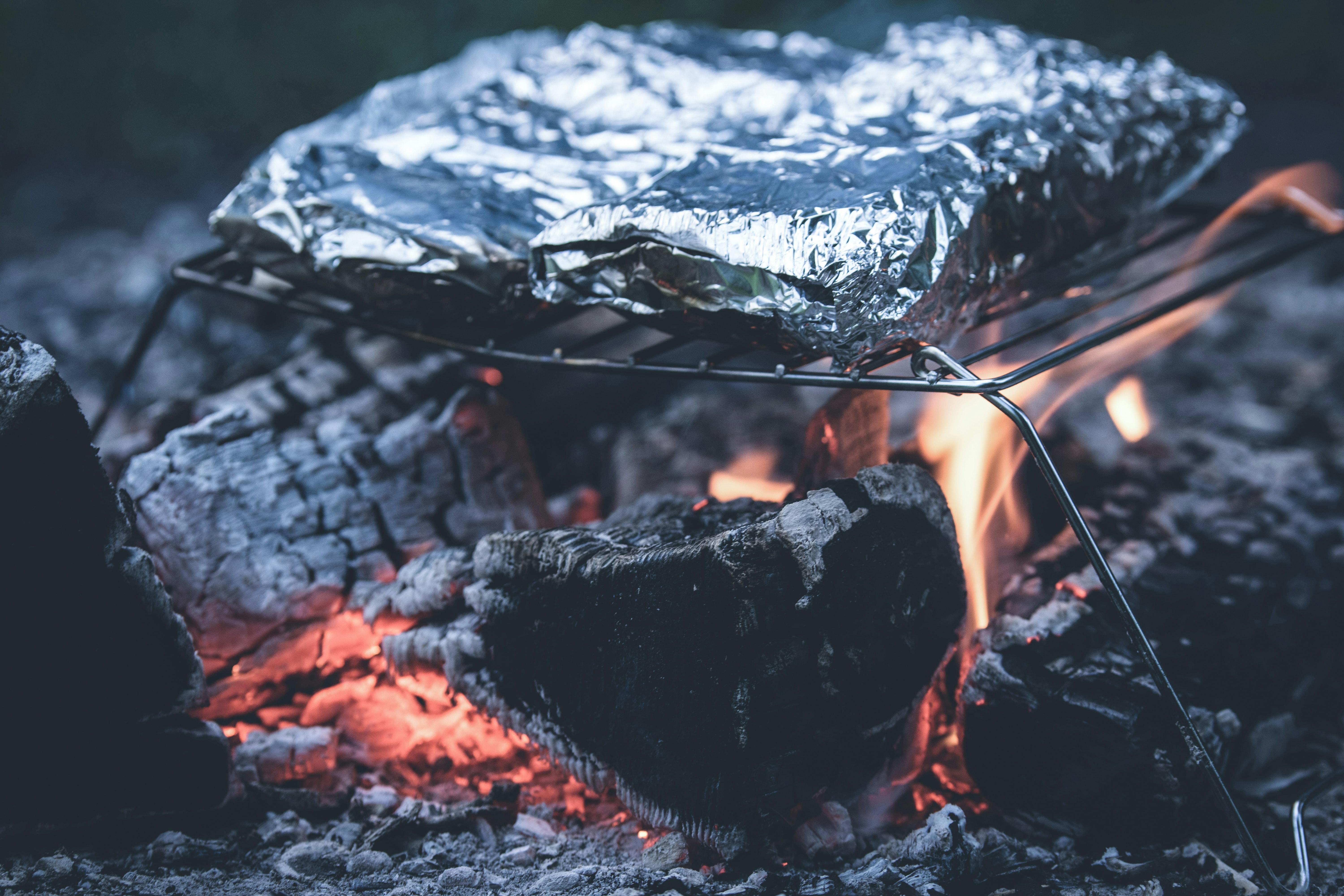 Something is wrapped in foil and placed on a grill above hot, smoldering logs and a small flame.