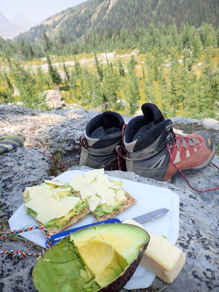 A pair of hiking boots and some avocado toast sit together on a rock in front of a forest