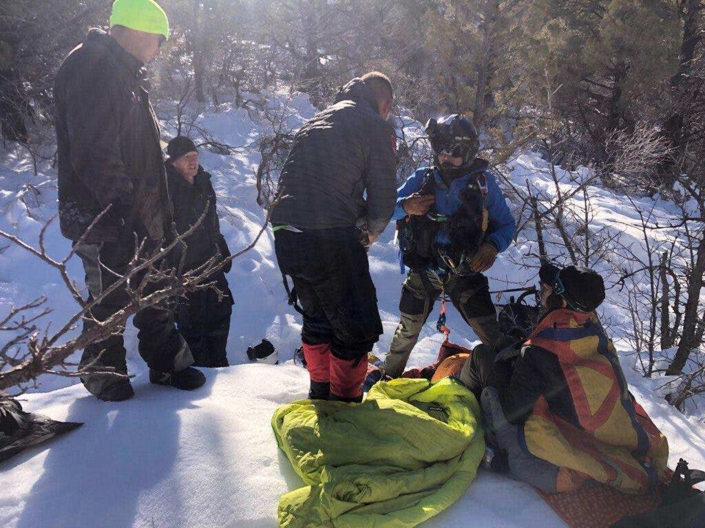 A search and rescue crew saves a woman in a snowy field. There are five rescuers wrapping her in a blanket.