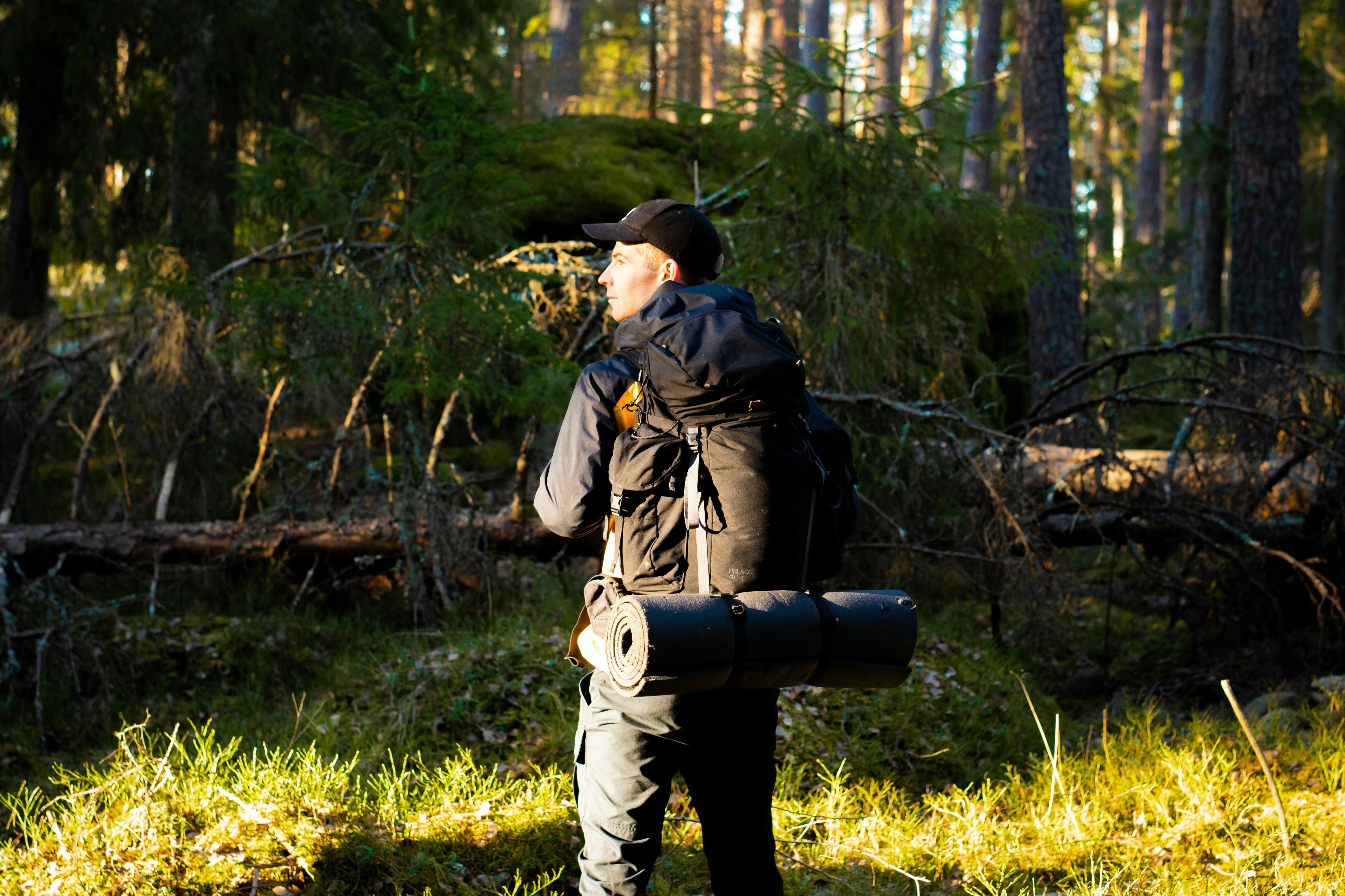 A man stands with his back to camera and his head turned sideways so his face is in profile. Ahead of him is a downed tree and sunlit grasses. He wears a large backpack with a rolled sleeping pad attached. 