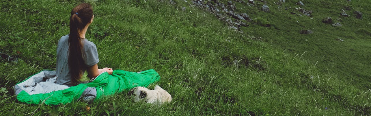 A girl in a green sleeping bag looks towards the distance while a pug sleeps next to her