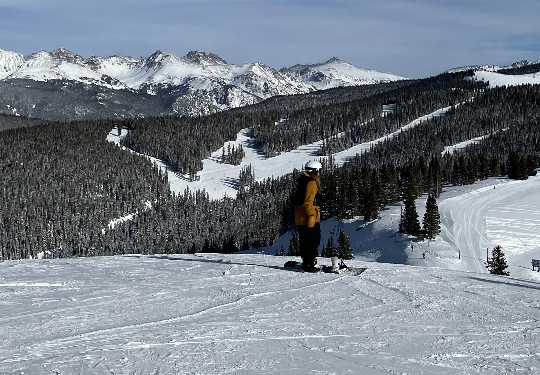 A man on a snowboard stands at the top of a ski run. There are mountains in the distance.