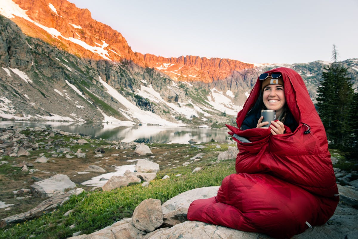The author sits on a boulder in her red sleeping bag, holding a warm mug. In the background is an alpine lake and peaks turned orange in the sunset/sunrise. 