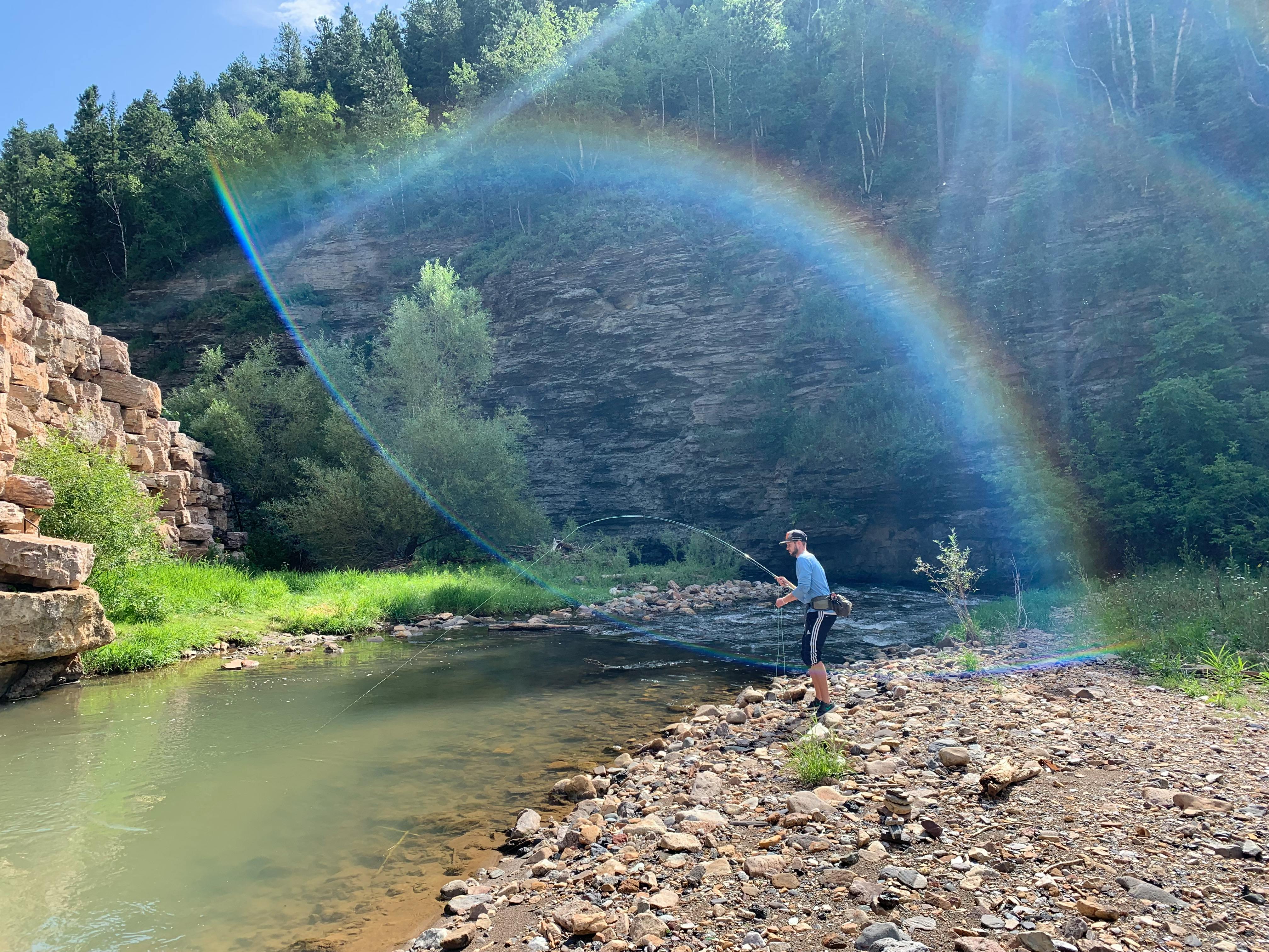 A man stands on the bank of a river and fly fishes. The banks are rocky and green and a rainbow from the lens curves over the scene.