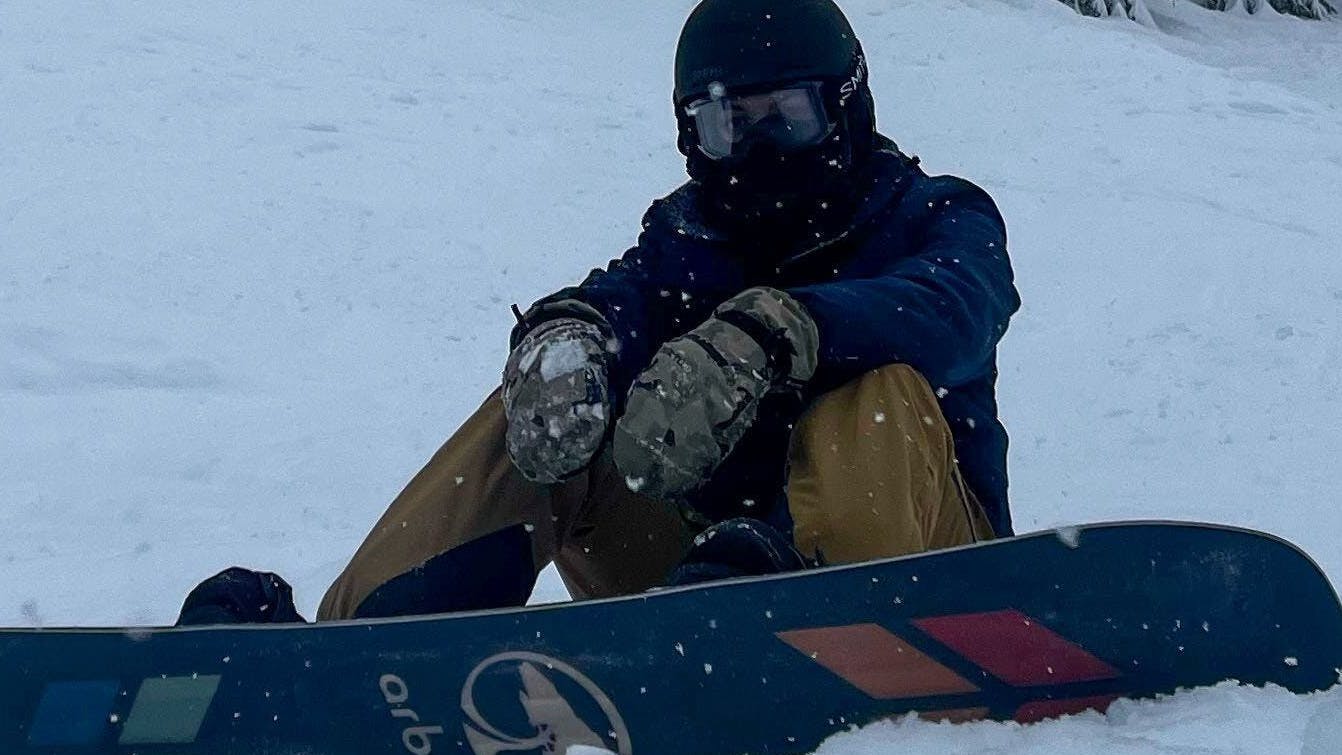 A snowboarder sitting on the Arbor Formula Camber snowboard. 