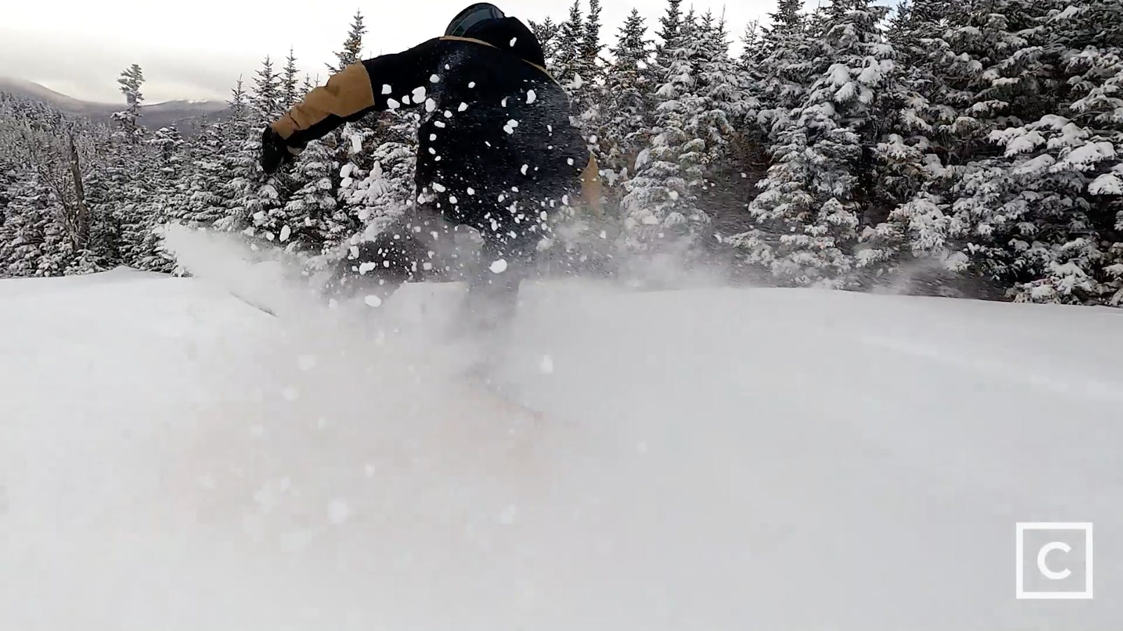 Curated expert Colby Henderson kicking up a spray of powder as he launches into a jump on his snowboard