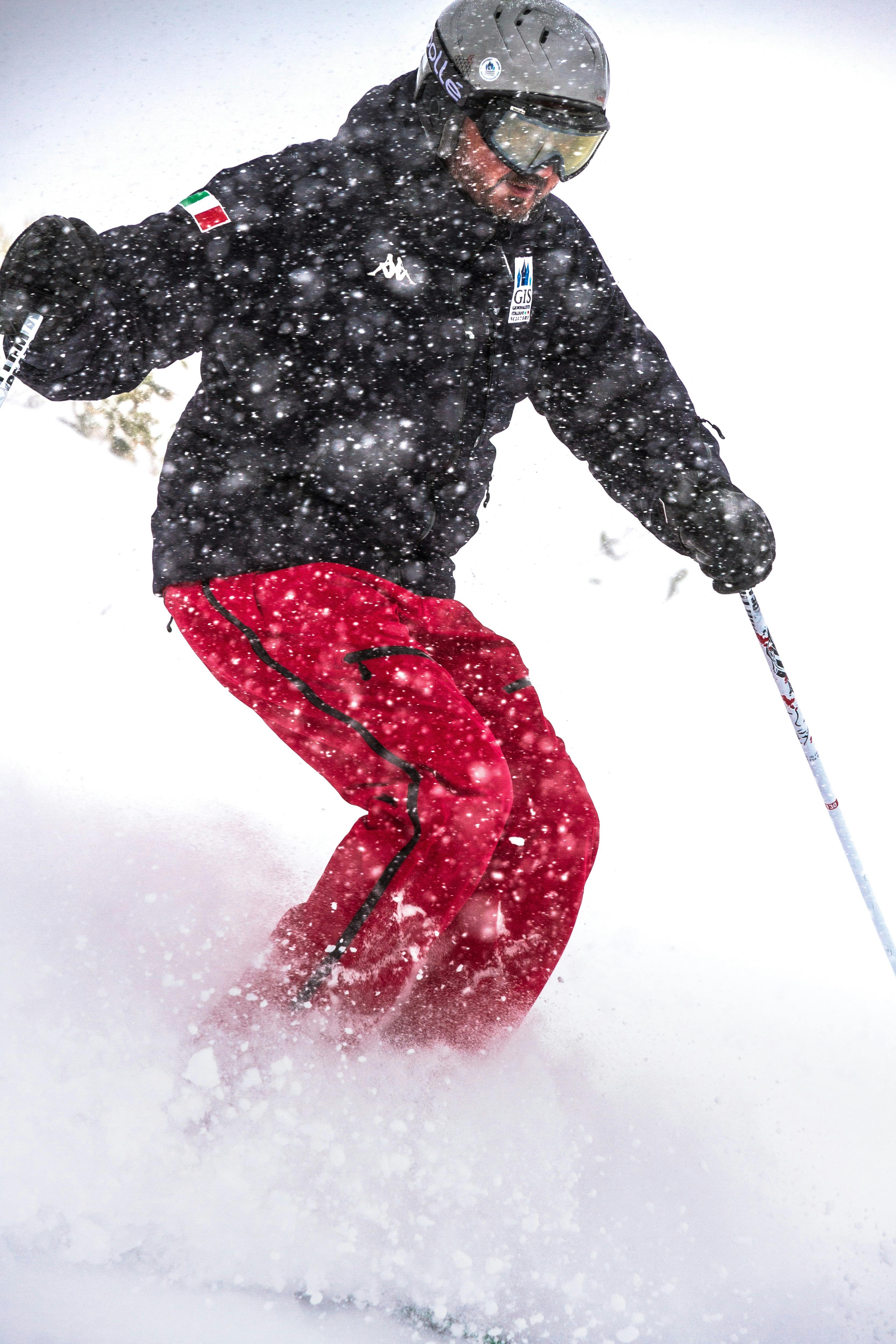 A close-up on a skier in a black jacket and red snow pants skiing down a slope, with knees deeply bent and kicking up lots of powder in the process.