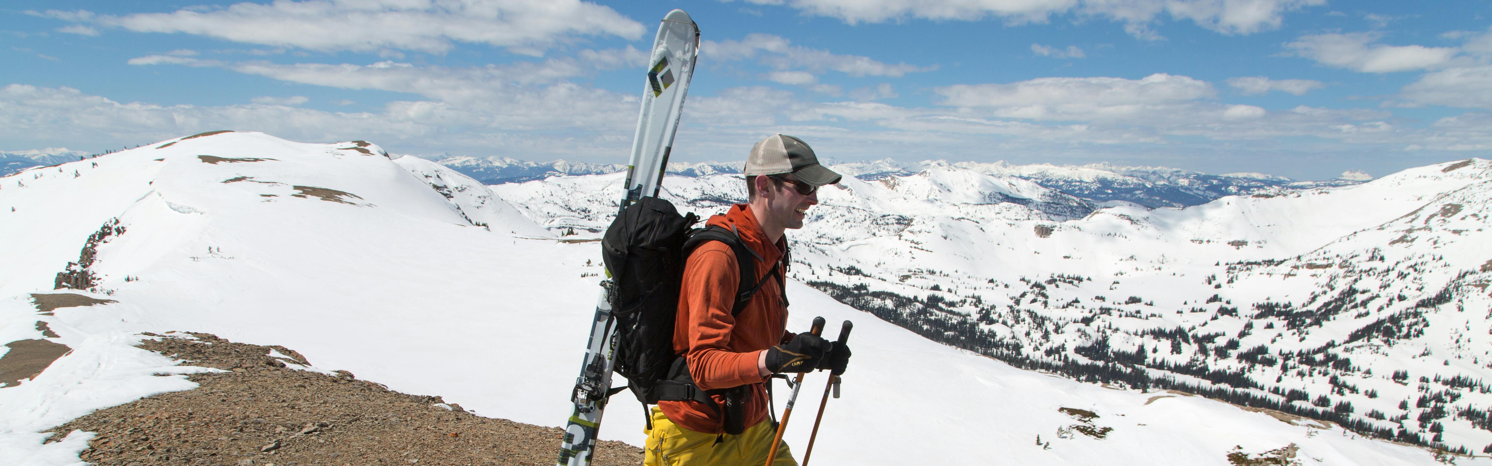A man walks on a ridgeline that is partly snowy and partly rocky. He wears one light layer and carries his skis on his backpack. 