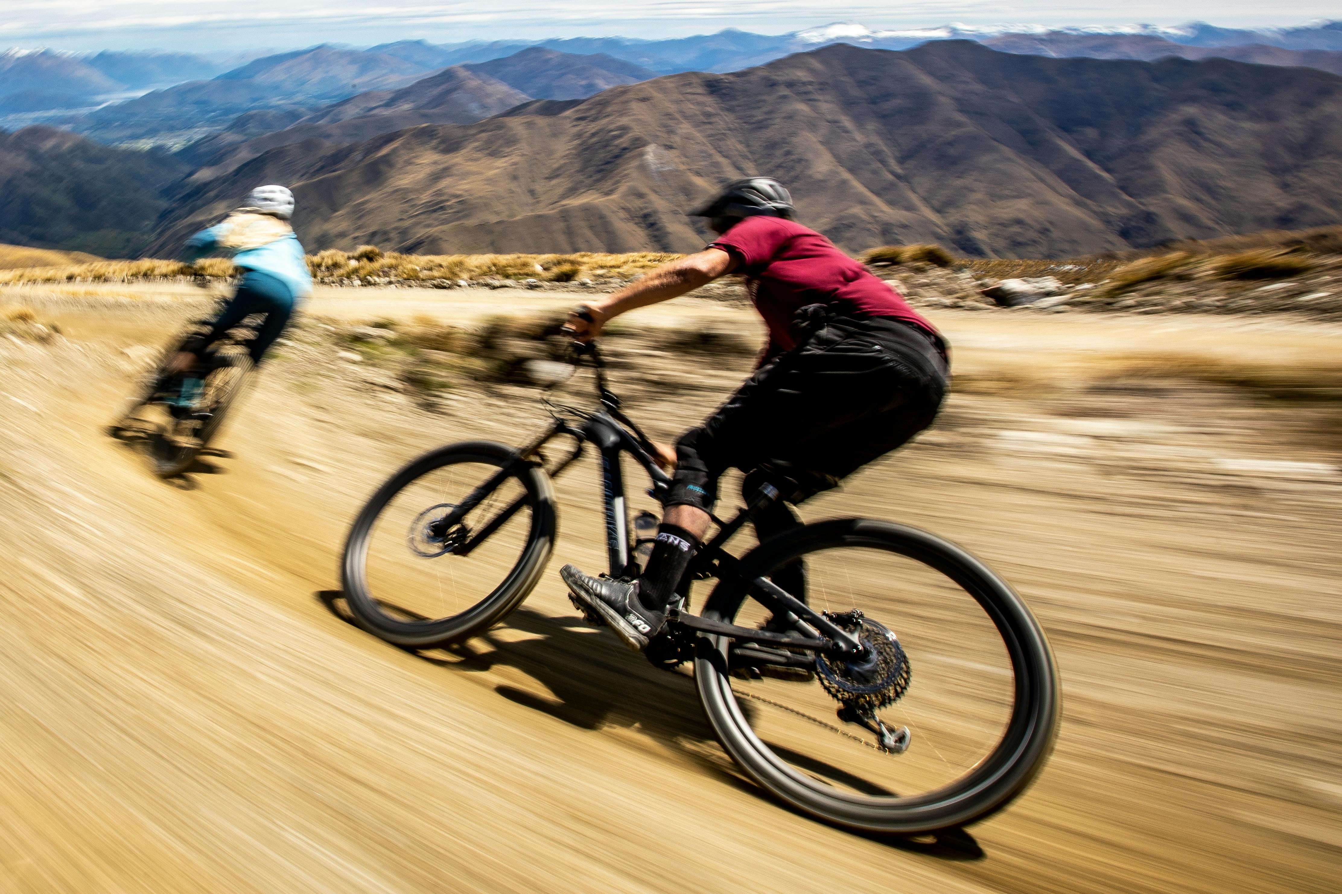 A man and woman speeding down a trail on their mountain bikes, mountain scenery in the background.