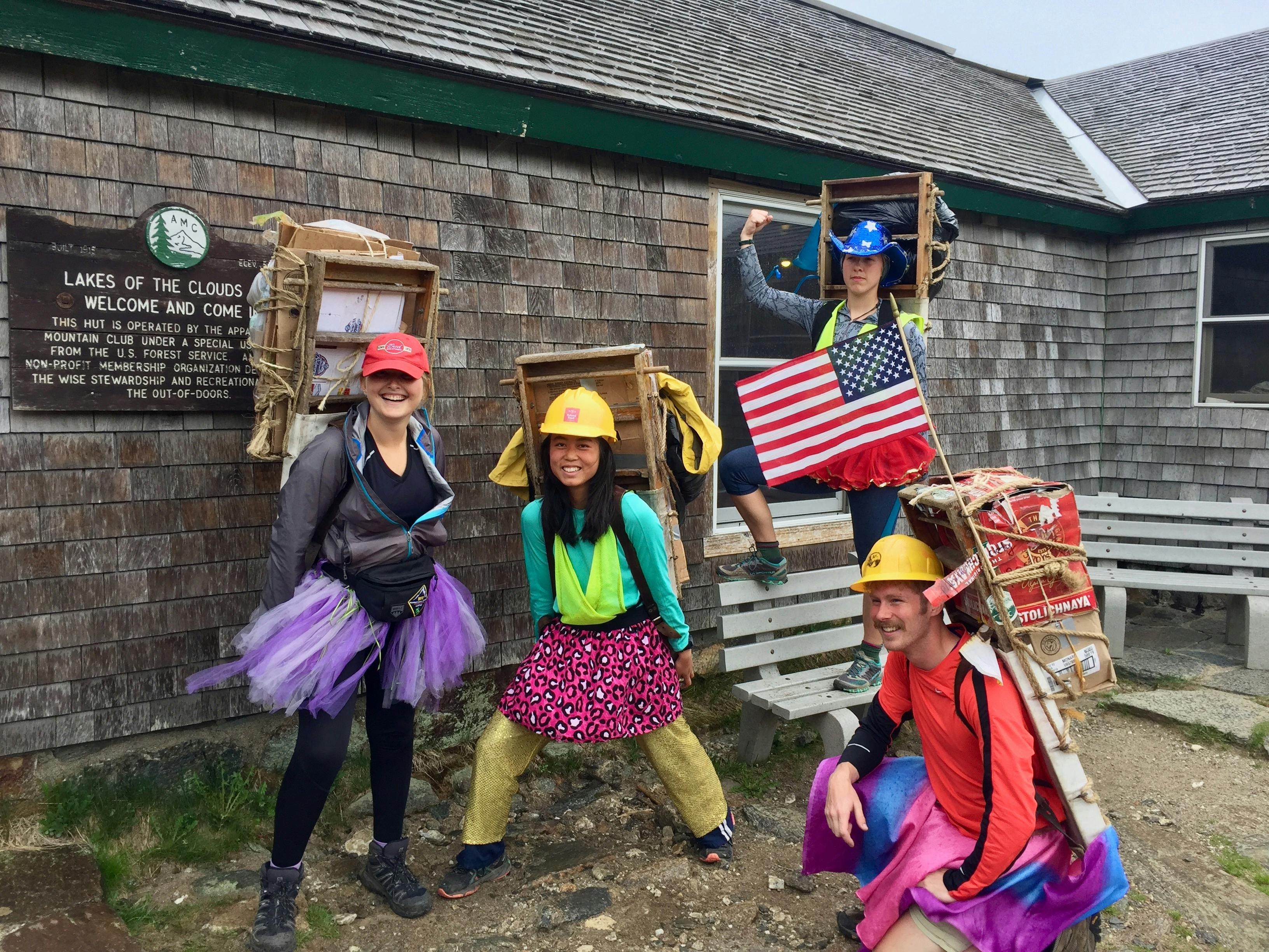 A group of people in silly skirts and costumes pose outside of a mountain hut.