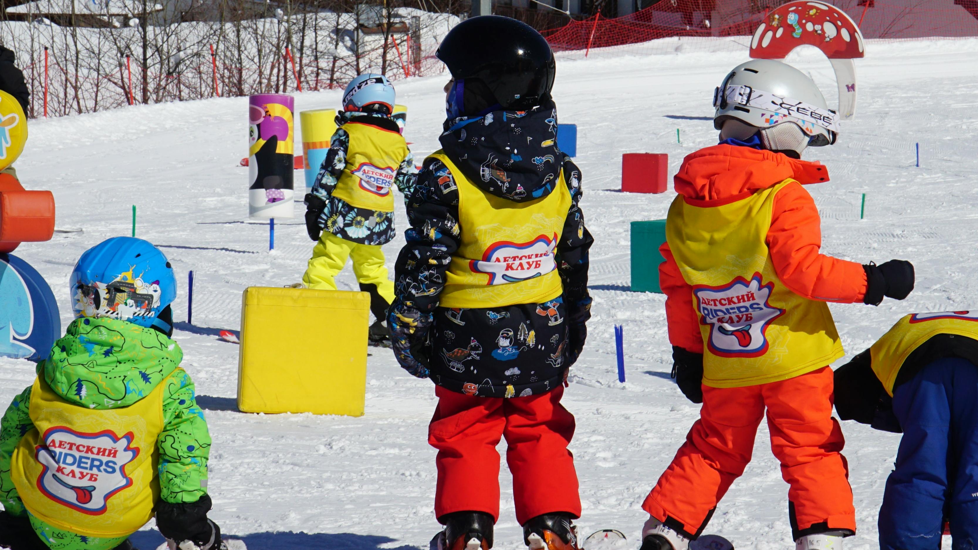 Several children are on snowboards in an obstacle course. They are all wearing the same shirt over their jacket and look like they are learning. There is an adult aged teacher standing in the background who looks like he is helping them learn how to snowboard. 
