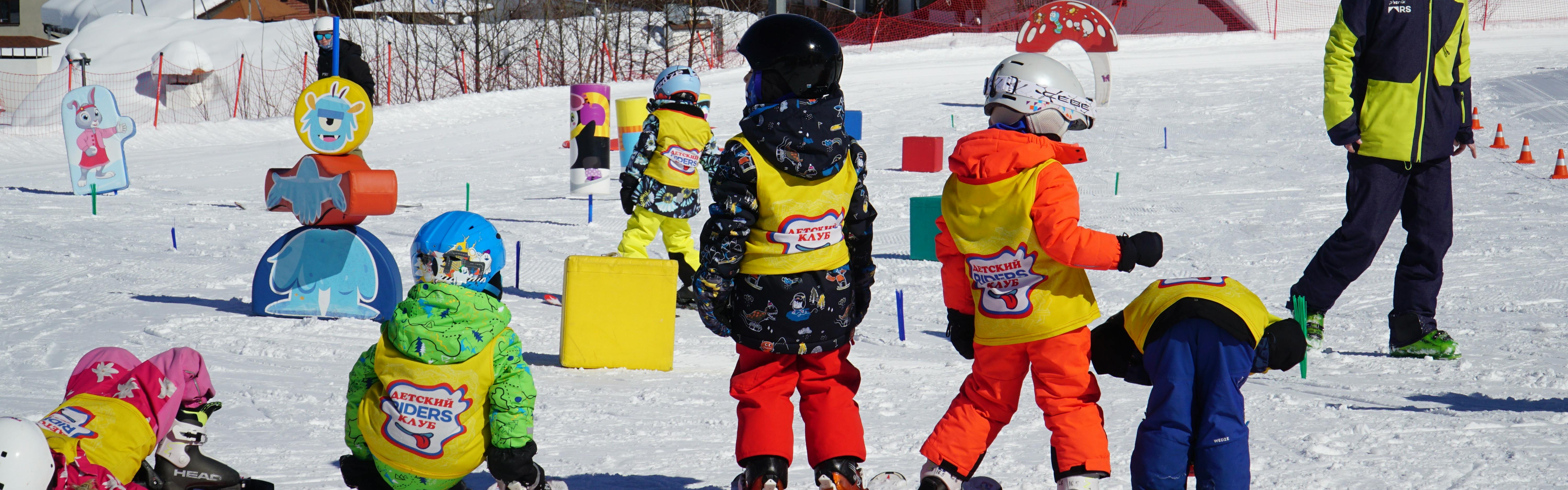 Several children are on snowboards in an obstacle course. They are all wearing the same shirt over their jacket and look like they are learning. There is an adult aged teacher standing in the background who looks like he is helping them learn how to snowboard. 