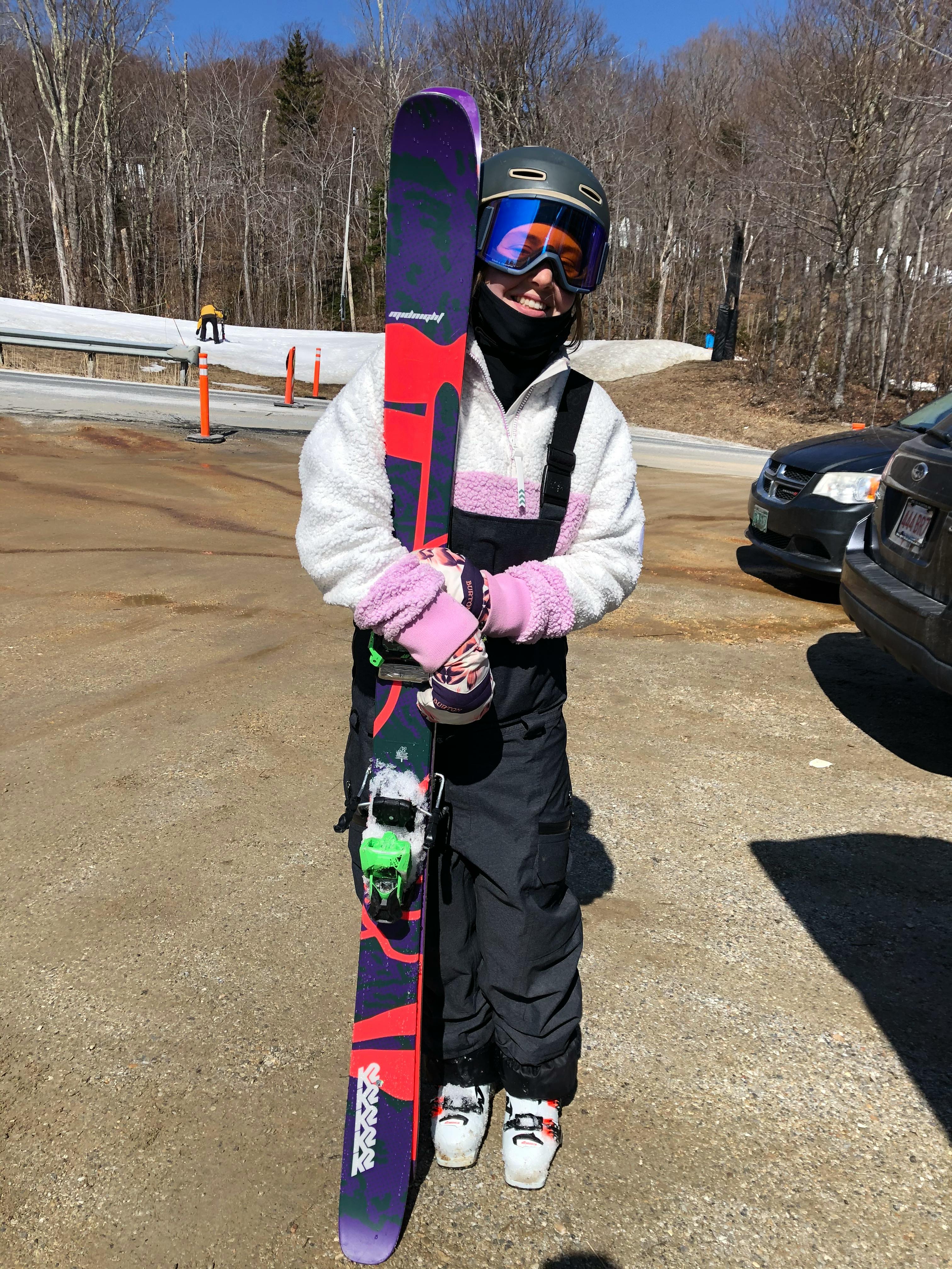 The author stands in the parking lot and smiles while holding her K2 skis. 