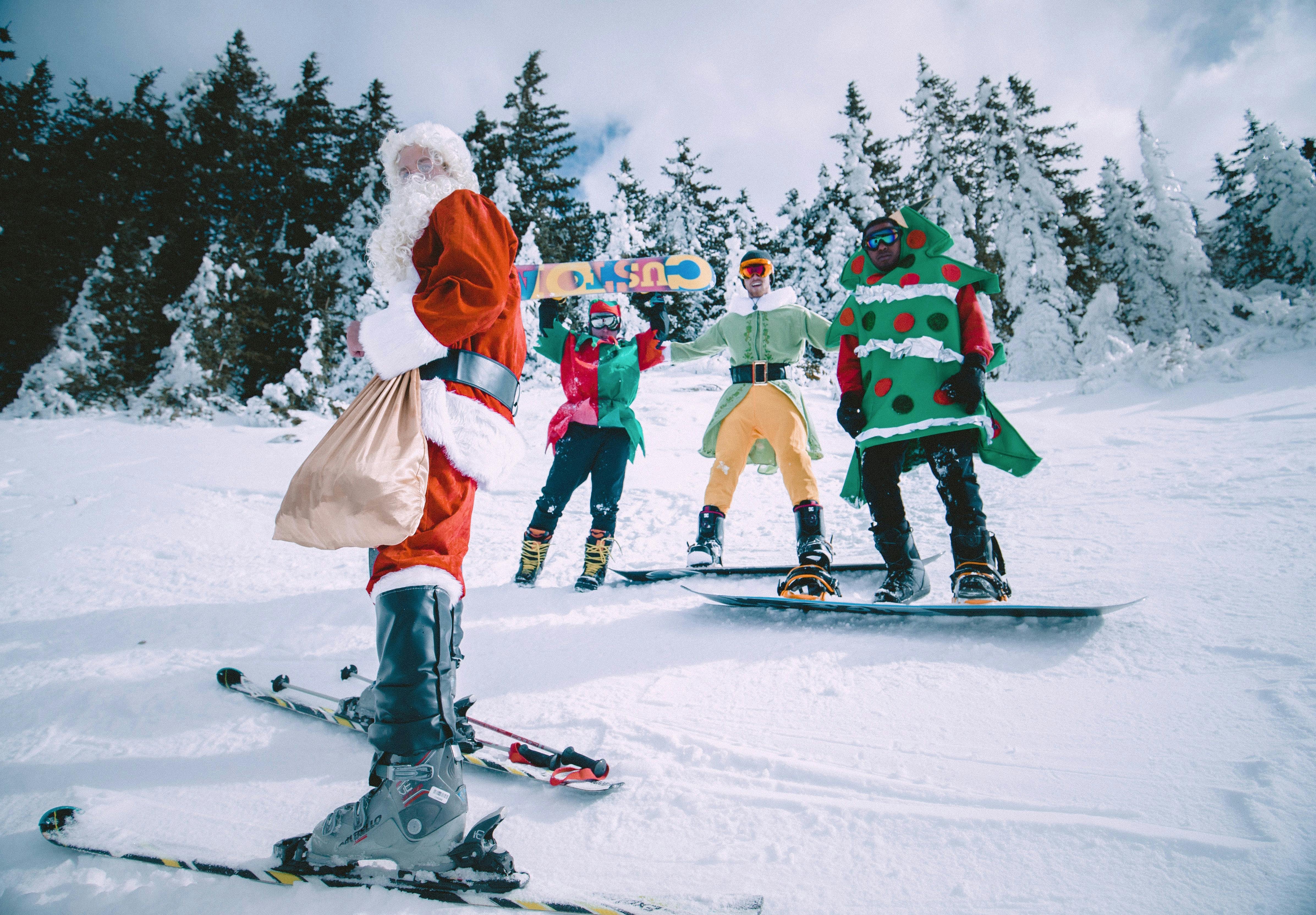A group of skiers and snowboarders stand together wearing Christmas costumes, including Santa, an elf, and a Christmas tree