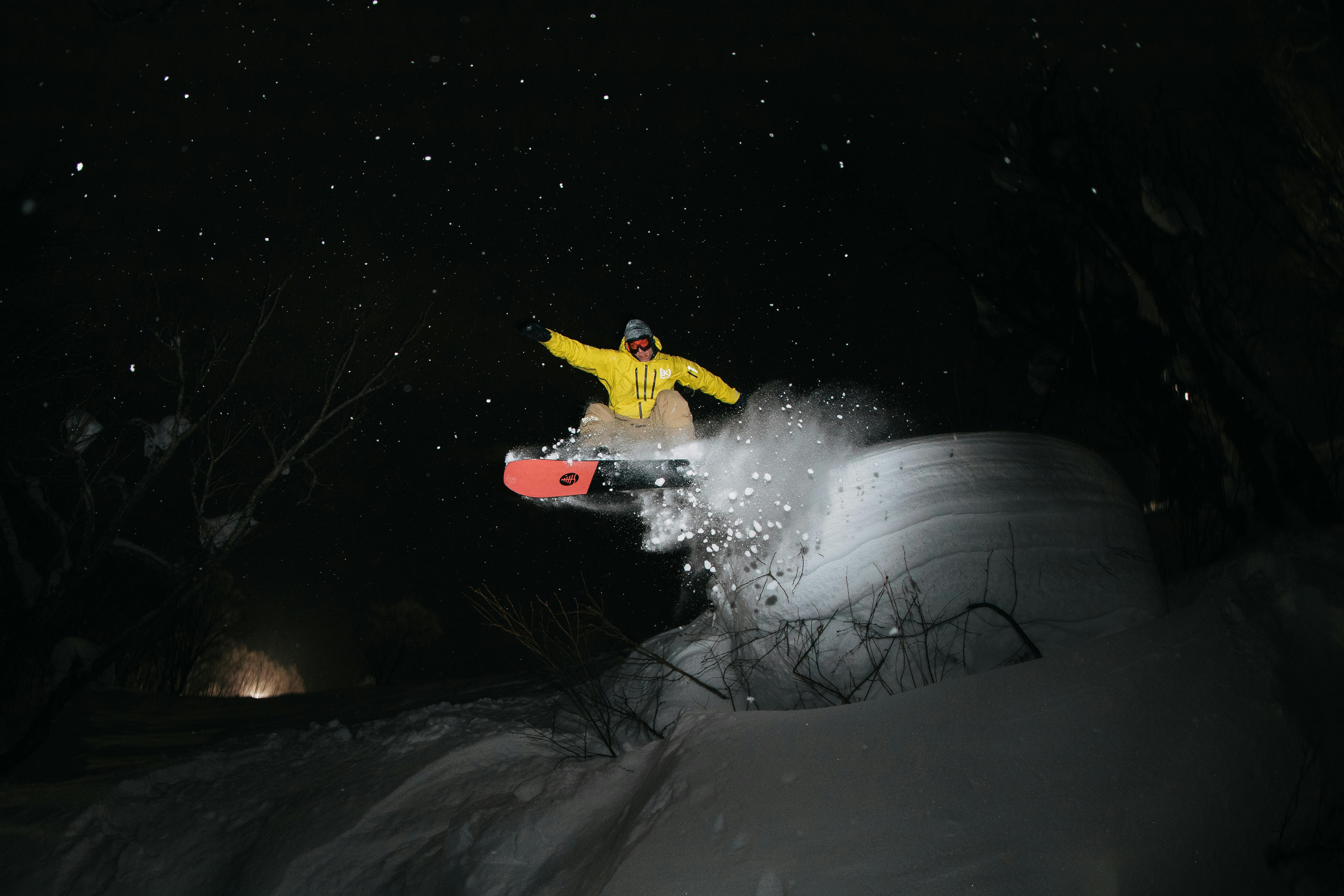 Dave catches air on his board at night, wearing a bright yellow jacket. The stars are visible in the night sky behind him.