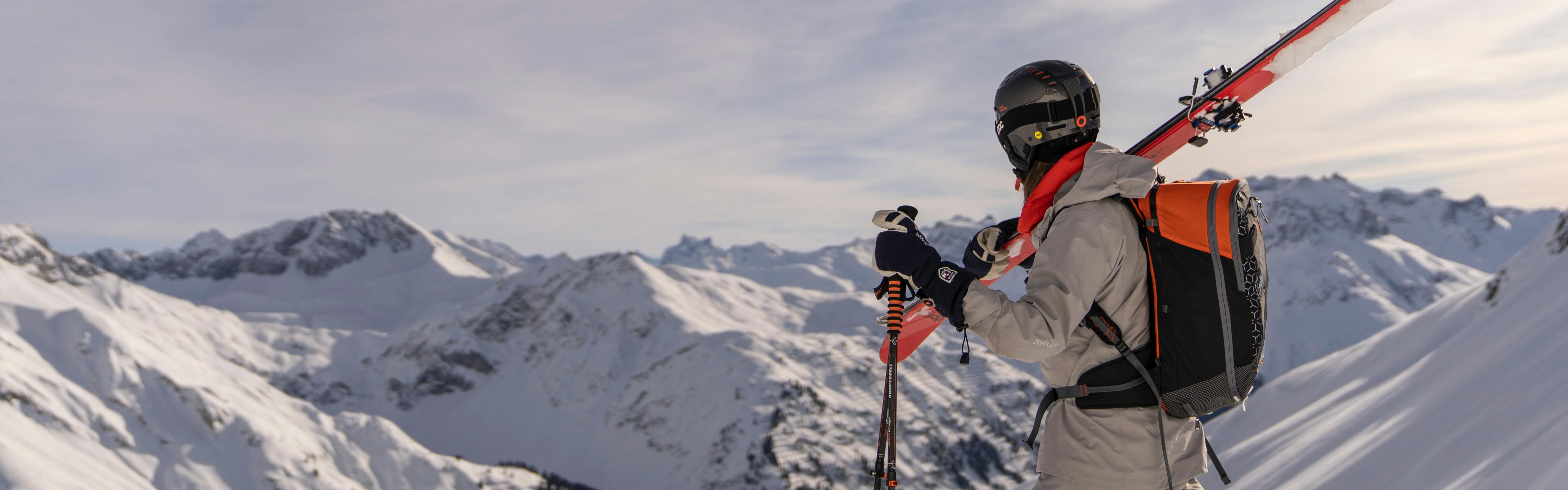 A skier stands on a peak and gazes off into the distance. They hold their poles in their hand, their skis over their shoulder, and wear a ski backpack.