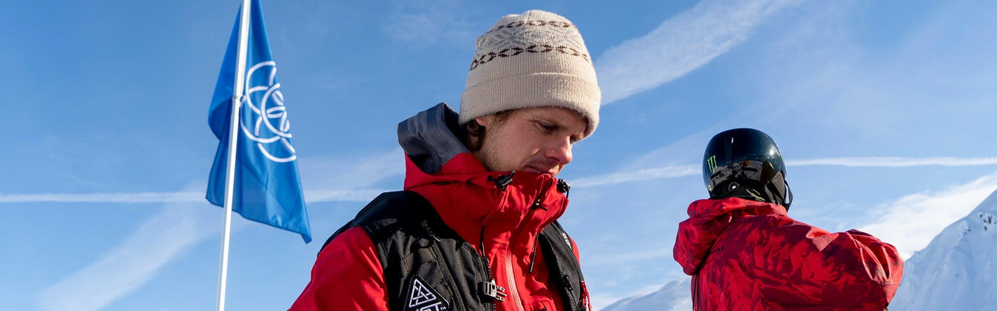 Snowboarder Dustin Craven stands in a red Natural Selection Tour jacket against a blue sky. 