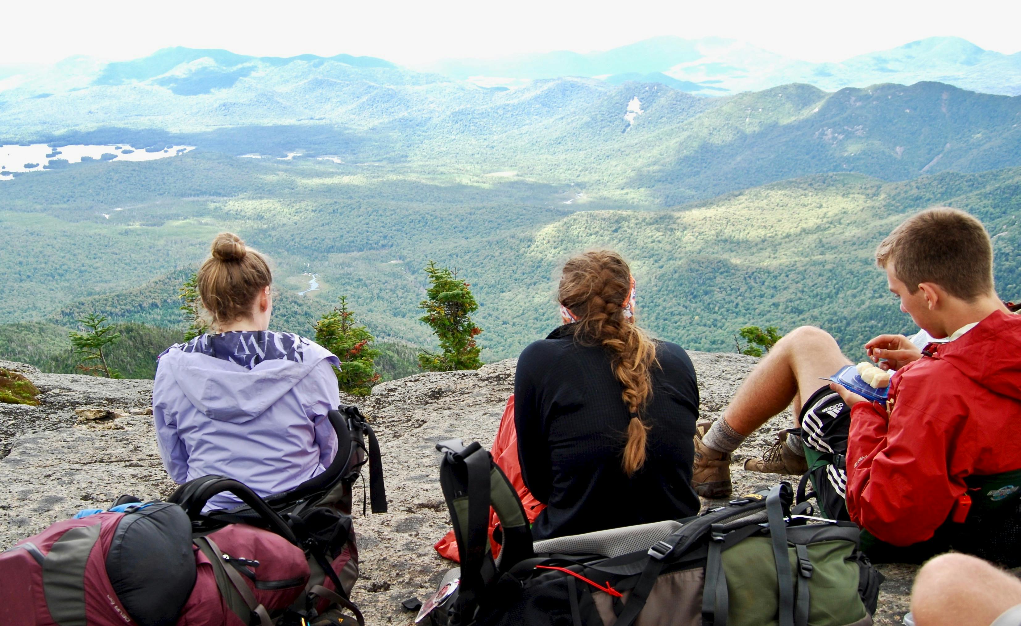Two women and a man sit on a rock with their backpacks on the ground behind them