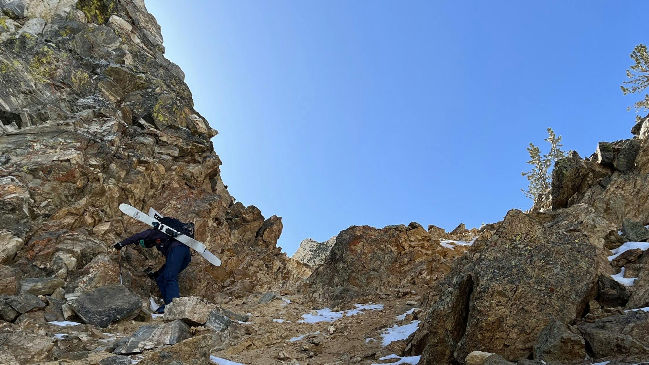 A woman with skis is climbing down a rocky face. She has skis on her backpack. 