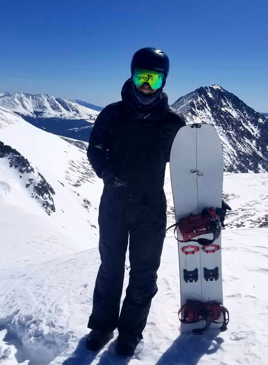 A man wearing snowboarding gear wearing the Giro MIPS helmet. He is holding a split-board and there are snowy mountains in the background. 